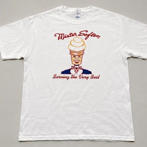 May include: White t-shirt featuring the Mister Softee logo. The logo depicts a smiling ice cream cone character in a blue suit and bow tie, with the text "Mister Softee" above and "Serving the Very Best" below.