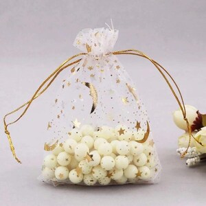 Peut inclure: Un sac en organza blanc transparent avec des &eacute;toiles dor&eacute;es et des croissants de lune imprim&eacute;s dessus. Le sac est rempli de perles blanches et nou&eacute; avec un ruban dor&eacute;.