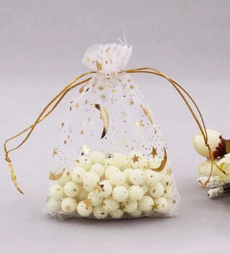 Peut inclure: Un sac en organza blanc transparent avec des &eacute;toiles dor&eacute;es et des croissants de lune imprim&eacute;s dessus. Le sac est rempli de perles blanches et nou&eacute; avec un ruban dor&eacute;.