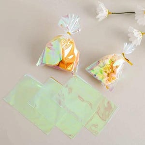 May include: Iridescent cellophane bags filled with treats, secured with gold twist ties. Several empty iridescent cellophane bags are also visible. The image is set against a neutral background, with artificial white flowers.