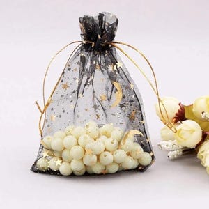Peut inclure: Un sac en tulle noir avec un cordon de serrage, imprim&eacute; d'&eacute;toiles dor&eacute;es et de croissants de lune. Le sac est rempli de perles blanches.