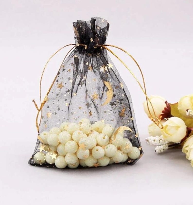 Peut inclure: Un sac en tulle noir avec un cordon de serrage, imprim&eacute; d'&eacute;toiles dor&eacute;es et de croissants de lune. Le sac est rempli de perles blanches.