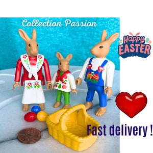 Può includere: Statuine a tema pasquale di coniglietti in abiti colorati, tra cui un vestito rosso e bianco, una camicia verde e bianca e una tuta blu. Sono visibili anche un cestino giallo, uova colorate e la scritta "Happy Easter".