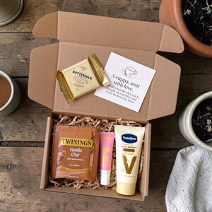 May include: A brown gift box containing a white chocolate bar, a Twinings Vanilla Chai tea bag, lip balm, and Vaseline hand cream. A card reads "A cuppa, sent with love." The box is lined with shredded paper.