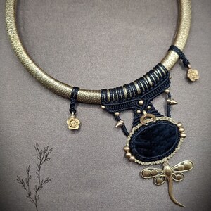 May include: A gold-toned collar necklace with a textured band and intricate black macrame detailing. The necklace features a dark blue oval pendant with a gold dragonfly charm and small gold accents.