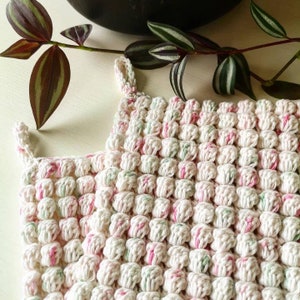 May include: A white crocheted pot holder with pink and green accents. The pot holder is made of a textured yarn and has a square shape.