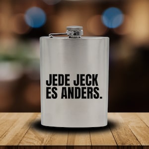 May include: A silver flask with a black inscription that reads "JEDE JECK ES ANDERS."