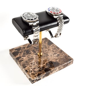 Puede incluir: Un expositor de relojes con dos relojes de pulsera. El expositor tiene una parte superior de polipiel negra, un poste de metal dorado y una base de mármol marrón. Los relojes tienen correas plateadas y esferas negras y azules.