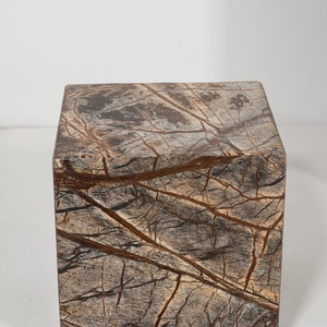 May include: A square, brown marble pedestal with a natural, veined pattern. The pedestal is made of a dark brown stone with lighter brown veins running through it.