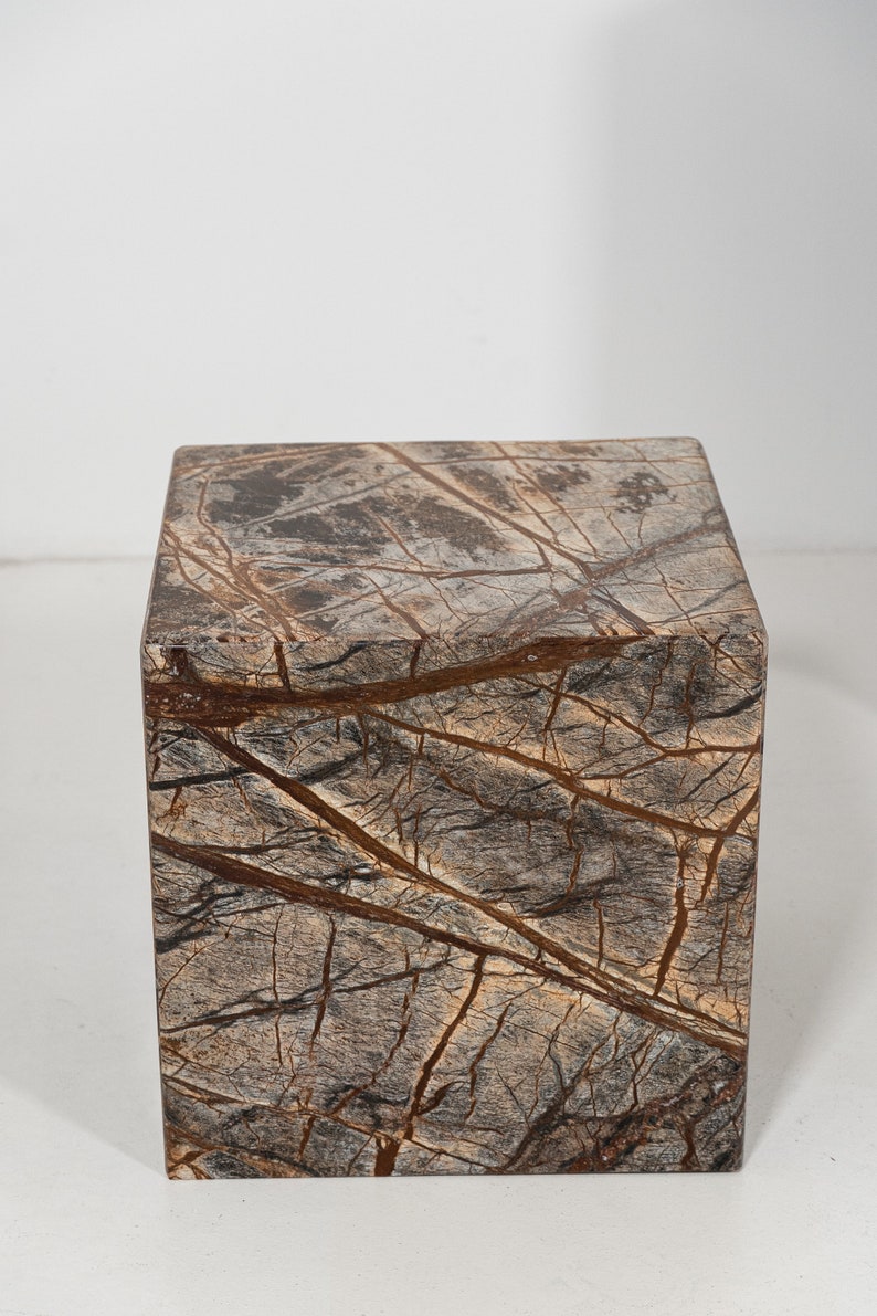 May include: A square, brown marble pedestal with a natural, veined pattern. The pedestal is made of a dark brown stone with lighter brown veins running through it.