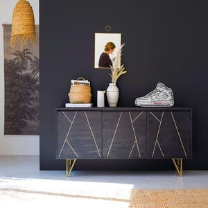 May include: A dark wood cabinet with gold accents and three doors. The cabinet has gold metal hairpin legs and sits on a light grey rug. A white sneaker is on top of the cabinet.