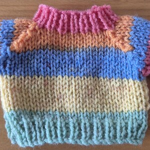 May include: A miniature, hand-knitted jumper in a vibrant array of colours. The jumper displays horizontal stripes of blue, yellow, orange, and green, with a pink collar and blue cuffs. The garment is made of soft yarn.