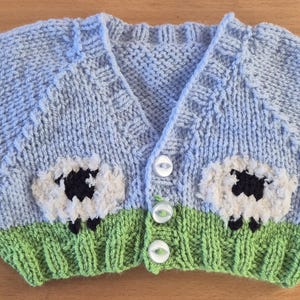 May include: A light blue knitted cardigan featuring a green ribbed hem and two white sheep motifs with black faces and legs. The garment has long sleeves, a V-neckline, and is fastened down the centre front with three white buttons.