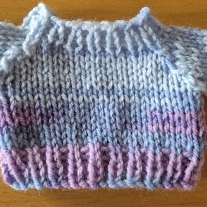 May include: A miniature hand-knitted sweater featuring a variegated yarn pattern in shades of light blue, lavender, and soft purple. The garment has long sleeves, a ribbed crew neckline, and a ribbed hem, constructed with a classic stockinette stitch.