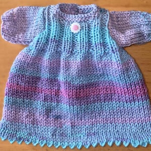 May include: A hand-knitted baby dress in shades of purple, blue, and pink. The dress features a scalloped hem, short sleeves, and a small flower-shaped button. The knitted pattern is visible throughout the garment.