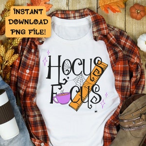 以下が含まれることがあります： 白いTシャツに黒い文字で「Hocus Focus」と書いてあります。テキストはクモの巣、クモ、紫色のコーヒーカップの定規で飾られています。