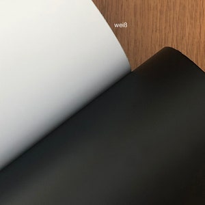 May include: A close-up of two sheets of paper, one white and one black, with the German words "wei&szlig;" and "schwarz" visible. The white sheet is on the left, and the black sheet is on the right. The background is a wood grain.