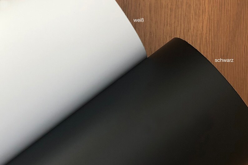 May include: A close-up of two sheets of paper, one white and one black, with the German words "wei&szlig;" and "schwarz" visible. The white sheet is on the left, and the black sheet is on the right. The background is a wood grain.
