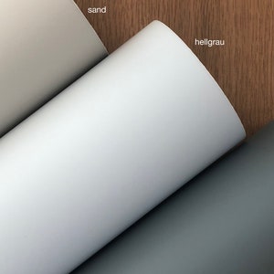 May include: Three rolls of paper in sand, light grey, and dark grey. The rolls are unrolled, revealing the different shades of colour. The background is a wooden surface.
