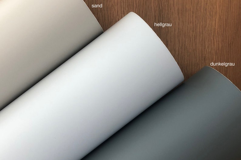 May include: Three rolls of paper in sand, light grey, and dark grey. The rolls are unrolled, revealing the different shades of colour. The background is a wooden surface.
