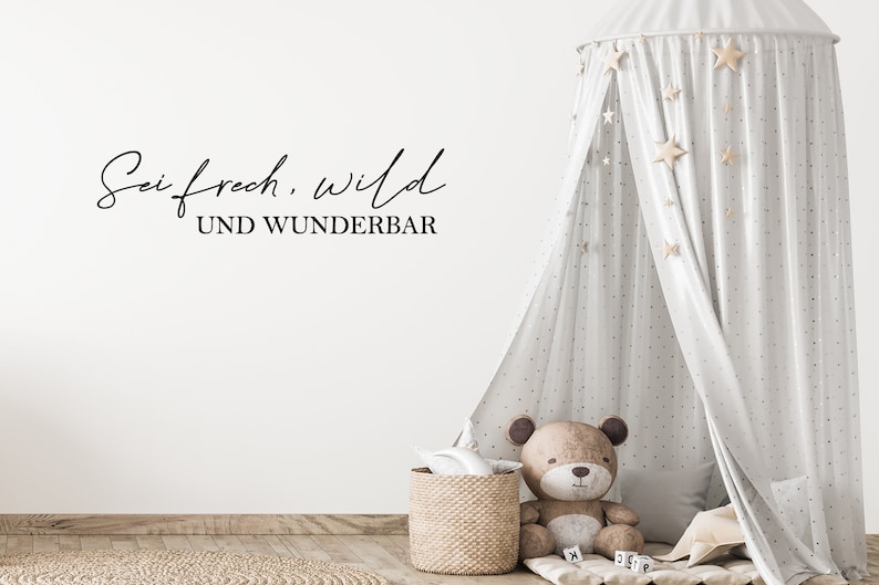 May include: A nursery scene featuring a light grey canopy bed with star decorations. A teddy bear sits beside a woven basket and a small rug. The wall displays the text "Sei frech, wild und wunderbar."