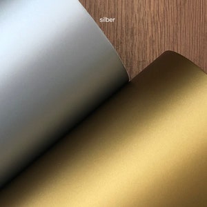 May include: Close-up of two sheets of paper, one silver and one gold. The silver sheet has the word "silber" printed on it, and the gold sheet has the word "gold" printed on it. The paper has a matte finish.