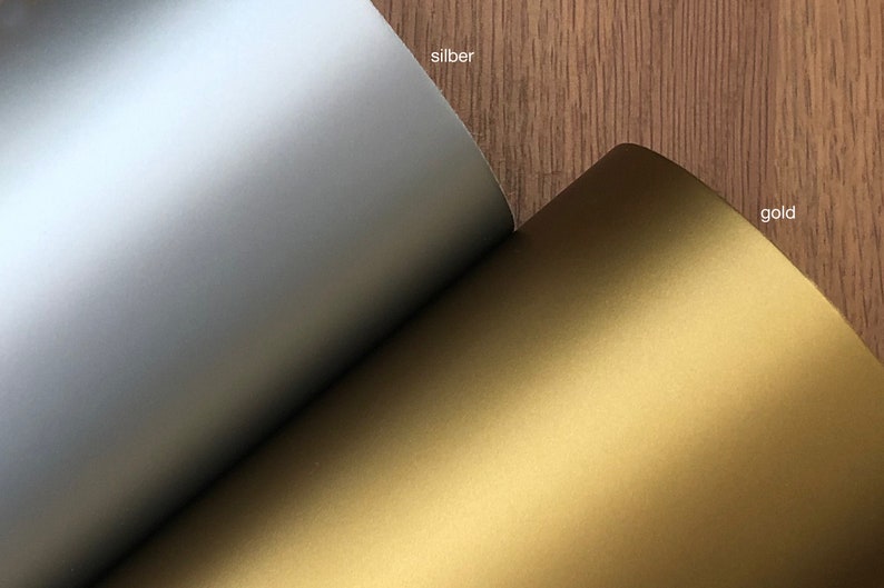 May include: Close-up of two sheets of paper, one silver and one gold. The silver sheet has the word "silber" printed on it, and the gold sheet has the word "gold" printed on it. The paper has a matte finish.