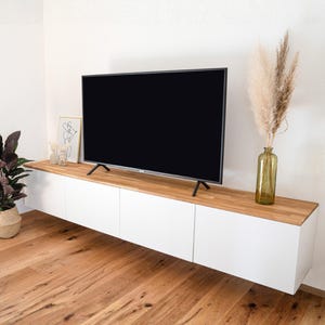 Könnte beinhalten: Moderne Wohnzimmereinrichtung mit einem großen schwarzen Flachbildfernseher auf einem schwebenden Schrank aus Weiß und Holz. Der Schrank steht an einer weißen Wand, mit einem Holzboden. Dekorative Elemente sind ein gerahmter Druck, eine Vase mit getrockneten Pflanzen und eine Topfpflanze.