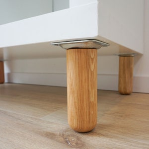 Könnte beinhalten: Ein weißer Schrank mit vier Holzbeinen. Die Beine sind mit Metallwinkeln am Schrank befestigt. Der Schrank steht auf einem Holzboden.
