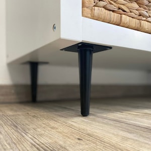 Könnte beinhalten: Ein weißer Schrank mit schwarzen, sich verjüngenden Beinen. Der Schrank hat eine Schublade aus geflochtenem Korb.
