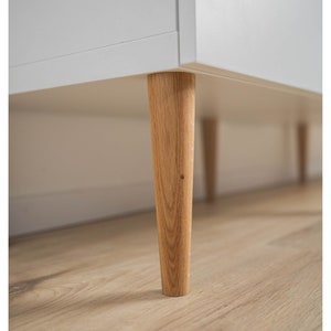 Könnte beinhalten: Nahaufnahme der Unterseite einer weißen Kommode mit vier Holzbeinen. Die Beine sind hellbraun und spitz zulaufend.