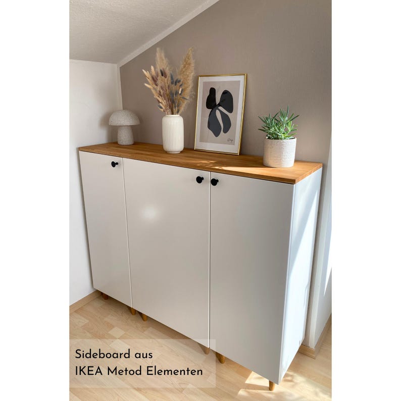 Beispiel für ein Projekt mit einer Massivholzdeckplatte nach Maß aus Eiche von Omformo. Hier als DIY Sideboard mit IKEA Metod Elementen, Möbelfüßen Upp in 10 cm Höhe und Möbelknöpfe Platta in Schwarz von Omformo. Dekoriert mit Pflanzen, einer weißen Vase mit Pampasgras und einer beigen Pilzlampe um ein Wohnbeispiel für einen modernes Sideboard darzustellen.