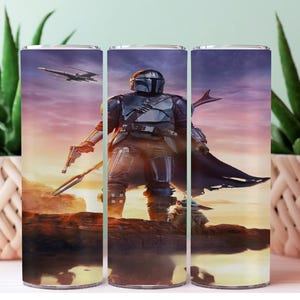 May include: A stainless steel tumbler featuring a detailed image of The Mandalorian from the Star Wars series. The design includes the character in armor, a blaster, and a spaceship against a sunset backdrop. The tumbler is cylindrical and likely holds approximately 20 ounces.