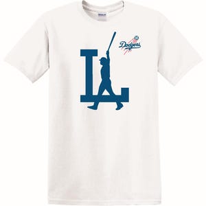 May include: White t-shirt with a blue silhouette of a baseball player holding a bat and the letters "LA" with the word "Dodgers" in blue text above the player.