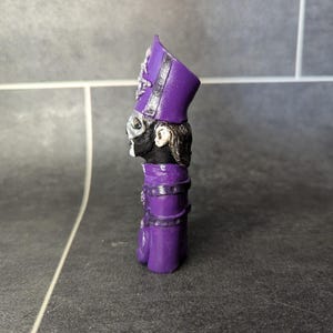 May include: A purple and black figurine of a person wearing a tall, purple hat with a black band. The figurine is standing on a gray surface.