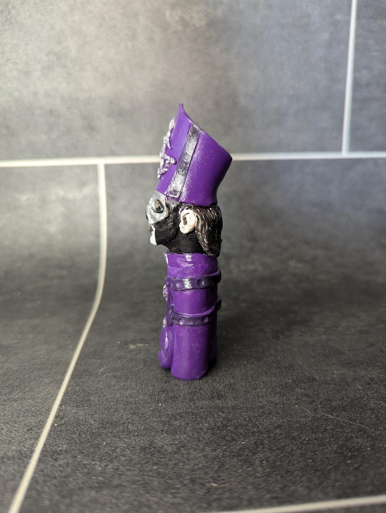 May include: A purple and black figurine of a person wearing a tall, purple hat with a black band. The figurine is standing on a gray surface.