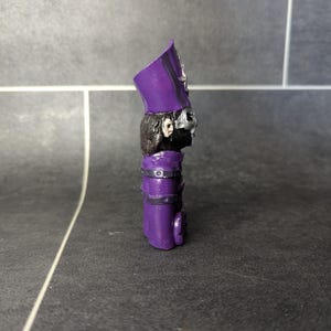 May include: Purple and black figurine of a character wearing a tall hat with a skull face mask.