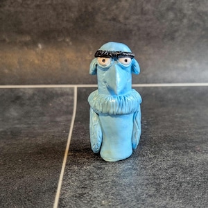 May include: A blue bird figurine with a black headband and angry eyes. The figurine is made of clay and has a textured surface.