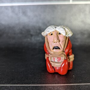 May include: A small, hand-crafted figurine of a person with white hair, a red and brown body, and a worried expression. The figurine is wearing a red and brown outfit with a number "13" on the front.