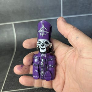 May include: A small purple figurine of a skeleton wearing a purple and silver bishop's hat with a skull and crossbones symbol. The figurine is holding its hands together in front of its chest.