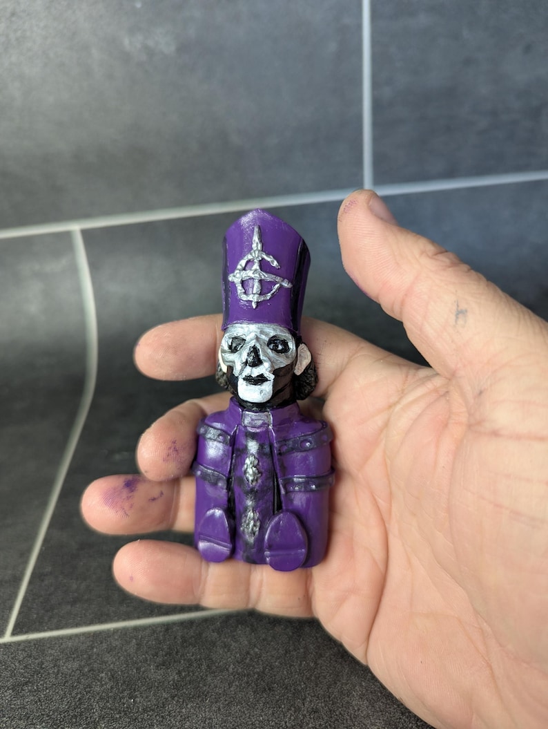 May include: A small purple figurine of a skeleton wearing a purple and silver bishop's hat with a skull and crossbones symbol. The figurine is holding its hands together in front of its chest.