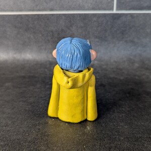 Puede incluir: Una peque&ntilde;a figura de un personaje con pelo azul y una chaqueta con capucha amarilla. La figura se ve desde atr&aacute;s, mostrando los detalles del atuendo y el peinado del personaje. La figura est&aacute; sobre un fondo oscuro.