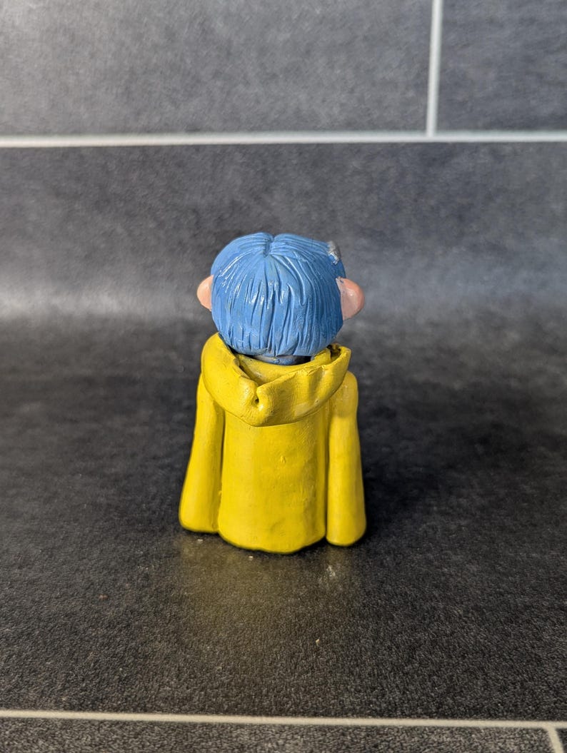 Puede incluir: Una peque&ntilde;a figura de un personaje con pelo azul y una chaqueta con capucha amarilla. La figura se ve desde atr&aacute;s, mostrando los detalles del atuendo y el peinado del personaje. La figura est&aacute; sobre un fondo oscuro.