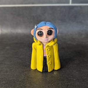 Puede incluir: Figura hecha a mano de un personaje con cabello azul, ojos de bot&oacute;n y un abrigo amarillo. La figura tiene una cara de color rosa claro y una peque&ntilde;a pinza plateada para el cabello. El abrigo tiene tres botones y est&aacute; abierto, revelando una camisa a rayas y pantalones negros.