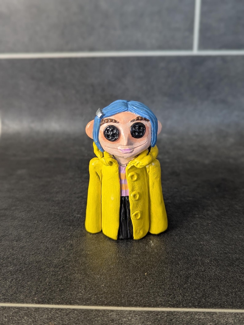 Puede incluir: Figura hecha a mano de un personaje con cabello azul, ojos de bot&oacute;n y un abrigo amarillo. La figura tiene una cara de color rosa claro y una peque&ntilde;a pinza plateada para el cabello. El abrigo tiene tres botones y est&aacute; abierto, revelando una camisa a rayas y pantalones negros.