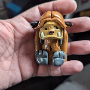 Puede incluir: Una pequeña figurita de arcilla marrón de una criatura con un cuerpo peludo, una cabeza grande y una sonrisa de dientes. La criatura tiene una cara amarilla con ojos negros y una nariz marrón. La figurita está sentada en cuclillas con las piernas cruzadas.