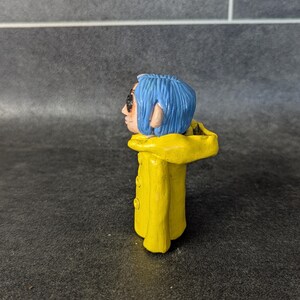 Puede incluir: Peque&ntilde;a figura hecha a mano de un personaje con cabello azul, gafas de sol y un impermeable amarillo. La figura tiene una cara detallada y est&aacute; de pie. El impermeable amarillo tiene tres botones visibles.