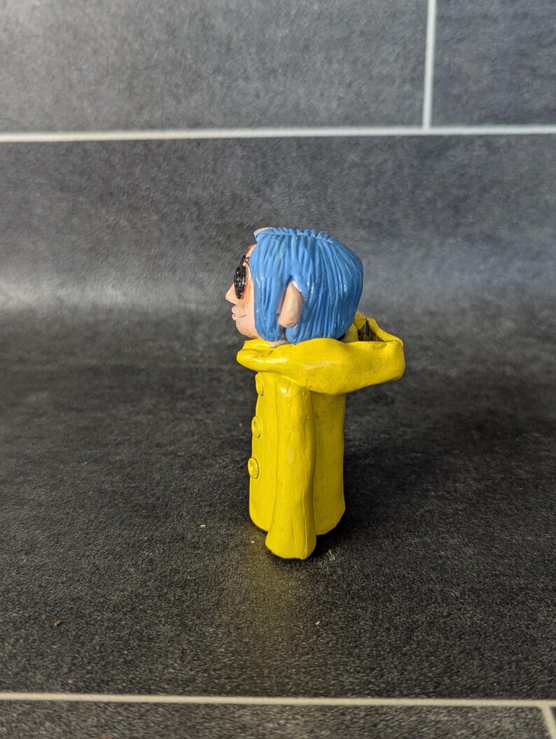Puede incluir: Peque&ntilde;a figura hecha a mano de un personaje con cabello azul, gafas de sol y un impermeable amarillo. La figura tiene una cara detallada y est&aacute; de pie. El impermeable amarillo tiene tres botones visibles.
