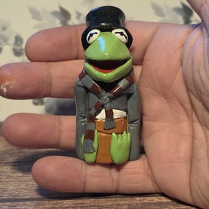 May include: A small green frog figurine wearing a black top hat, a grey coat, and a brown and grey striped scarf. The frog has a wide mouth, large eyes, and is positioned in a seated posture with its hands resting on its lap over a brown base.