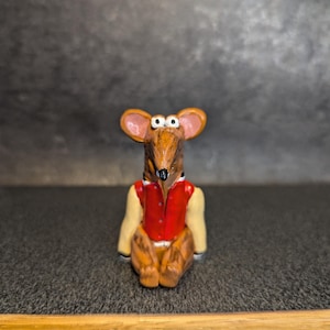 May include: A brown and tan ceramic mouse figurine wearing a red waistcoat and a white jacket. The mouse has large white eyes and is sitting with its legs crossed.